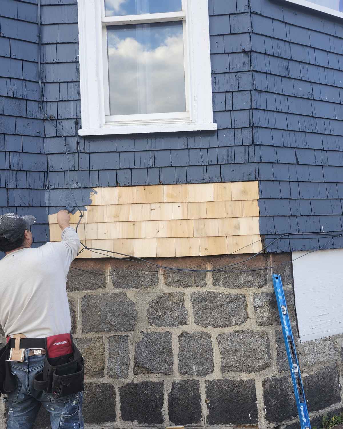 Roofer replacing damaged cedar shingle siding with new shingles above a stone foundation