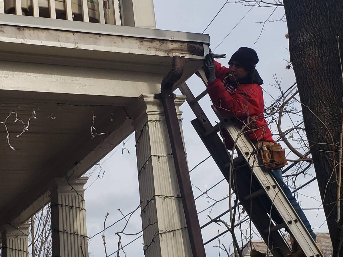 Roofer on a ladder installing a downspout on a house with a columned porch in winter