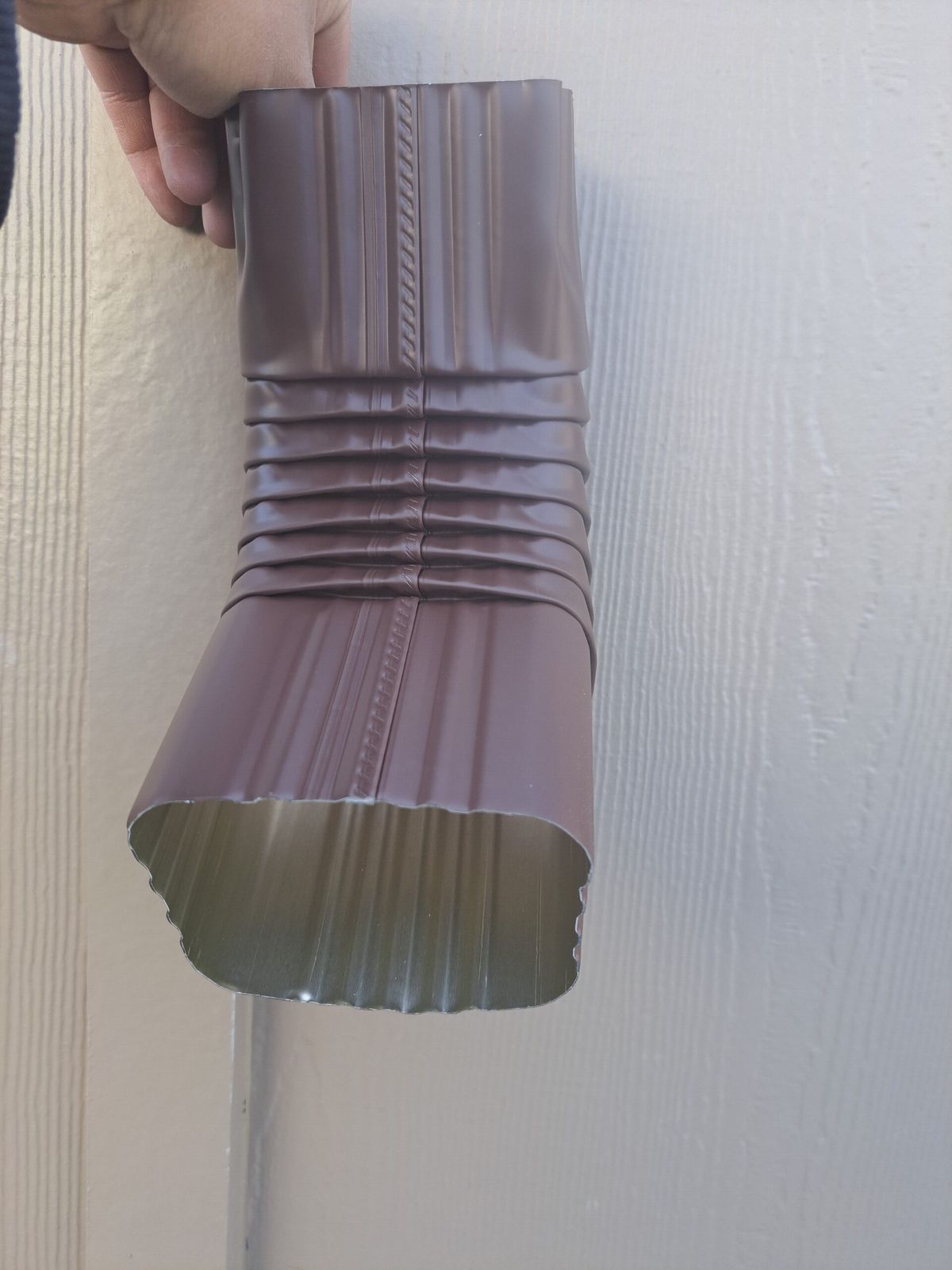 Hand holding a brown aluminum downspout elbow against siding