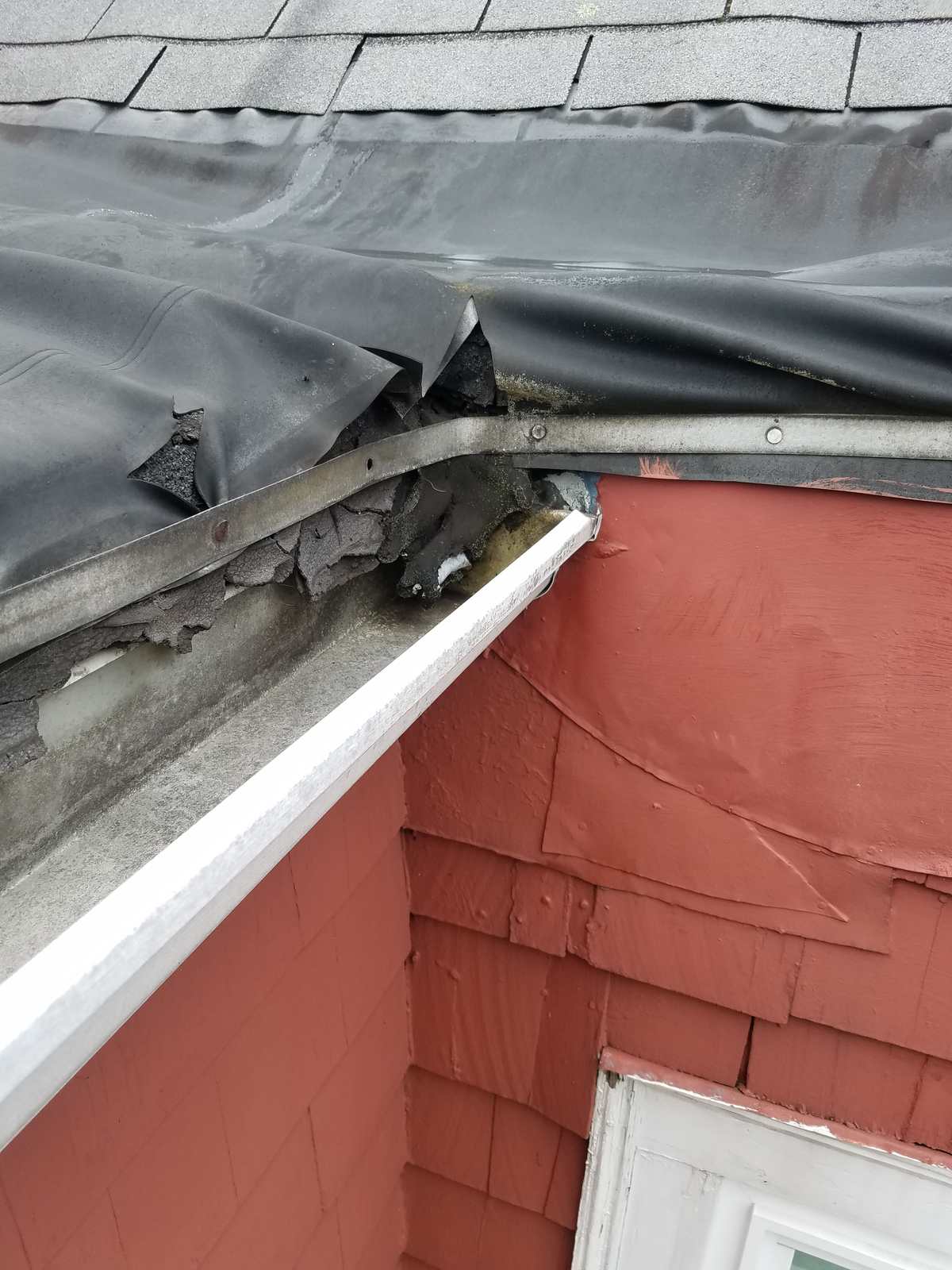 Damaged roof edge with torn rubber membrane and gutter at a red-shingled wall before repair
