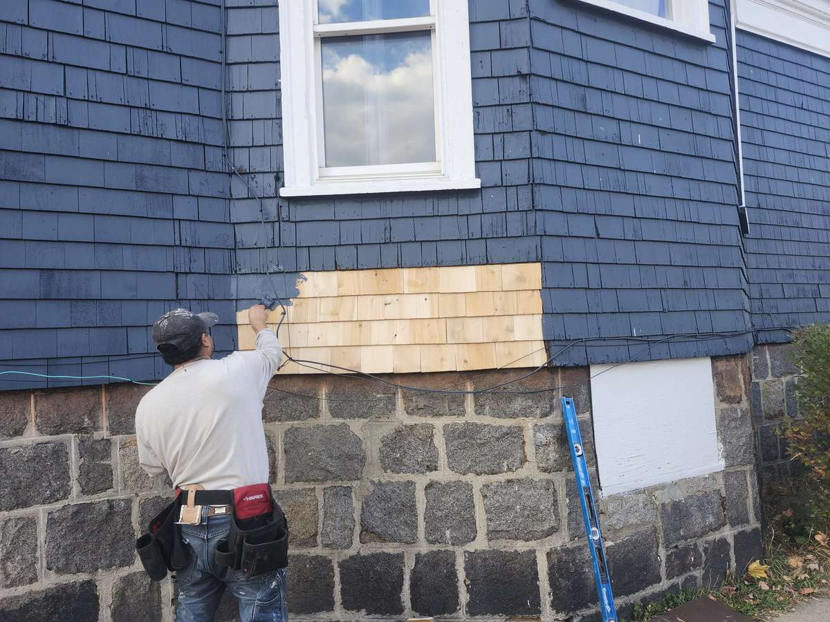 Roofer replacing damaged cedar shingle siding with new shingles above a stone foundation