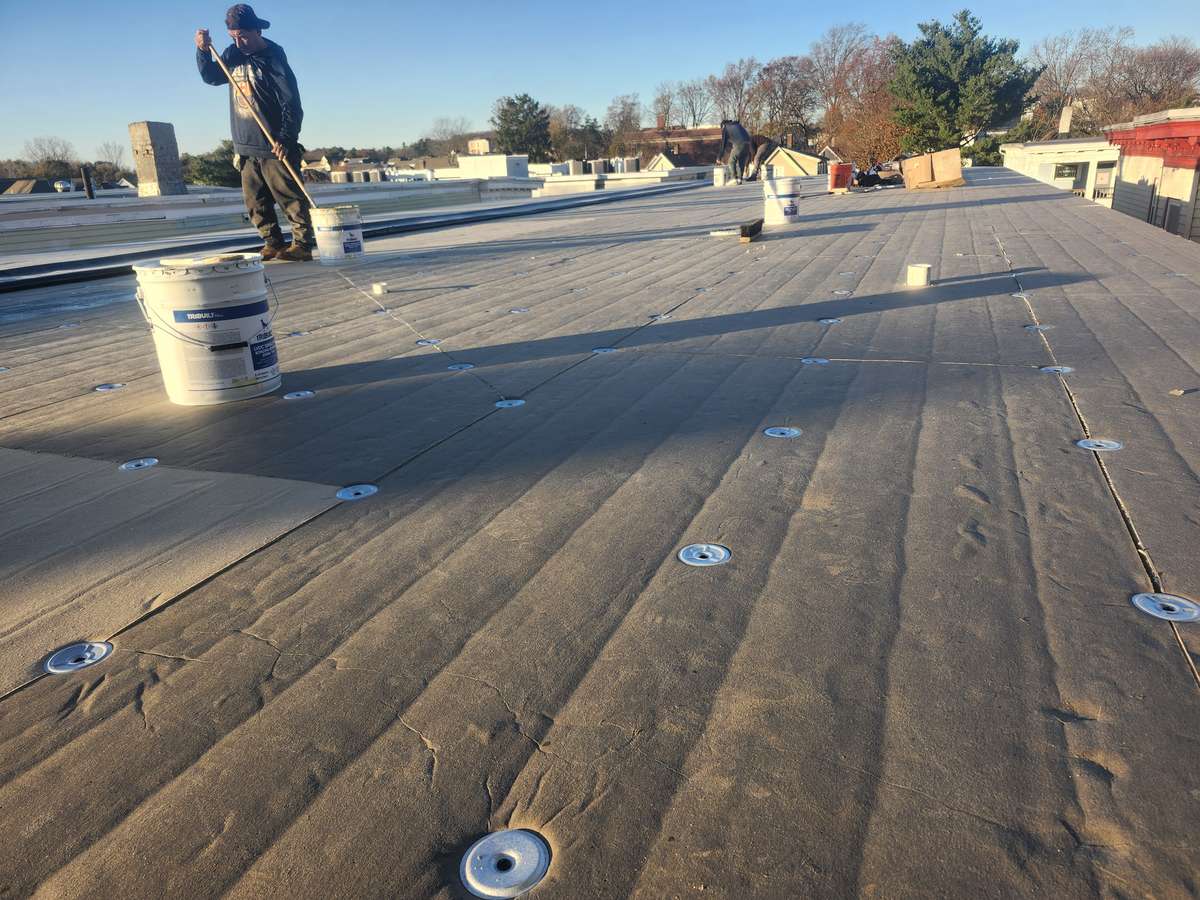 Roofer applying rubber roofing on a large flat roof with adhesive bucket and fastener plates