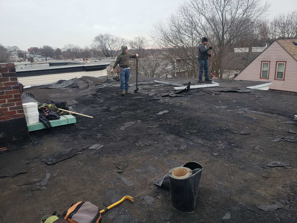 Roofers tearing off an old flat roof with debris and tools, houses behind