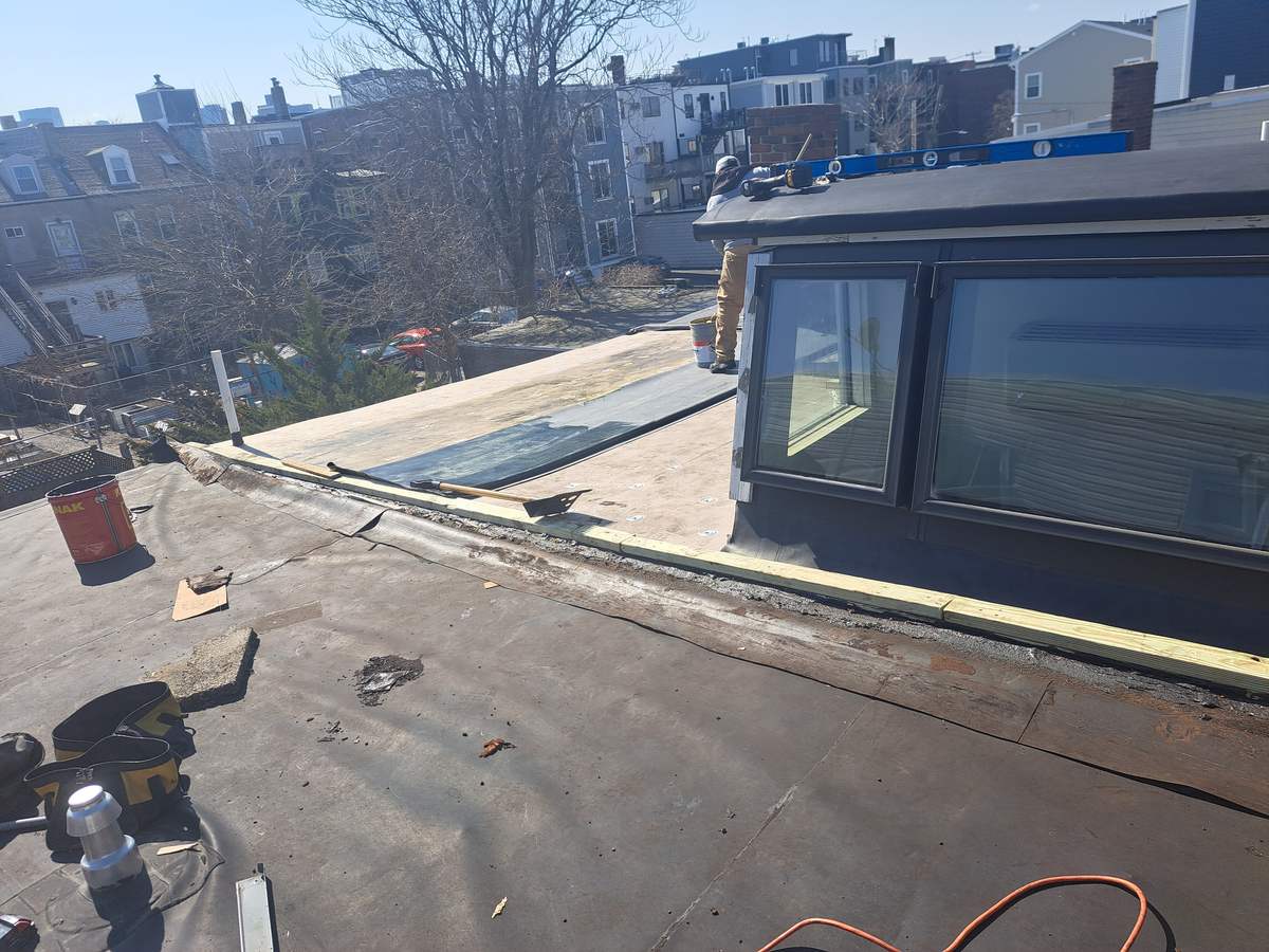 Flat roof under renovation with old decking exposed at the edge, city rooftops behind