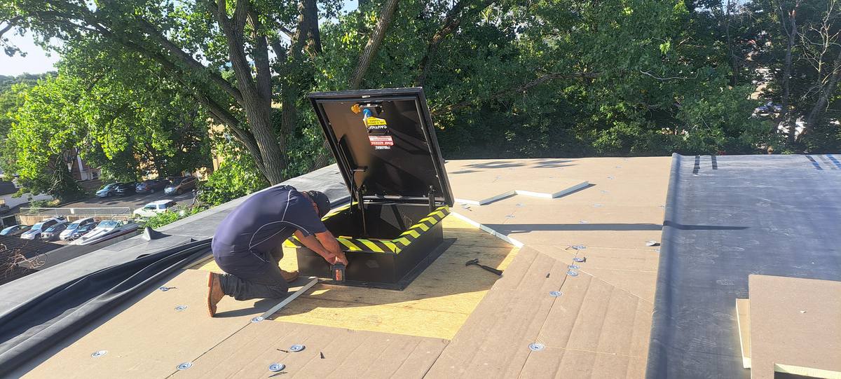 Roofer fastening insulation boards on a flat roof next to an open roof hatch