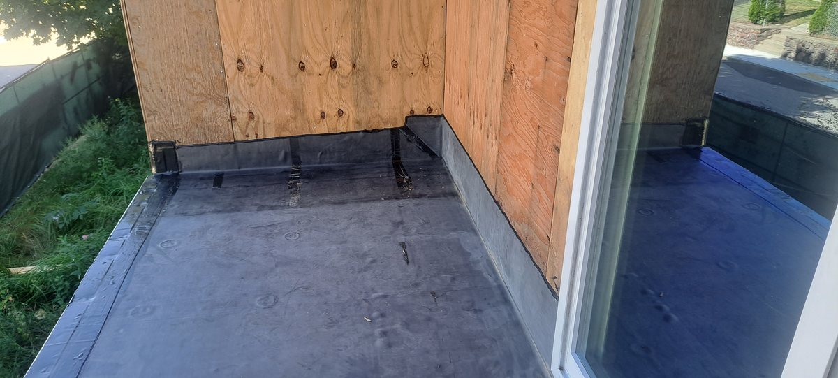 Rubber membrane at a flat roof corner with plywood parapet and white window trim
