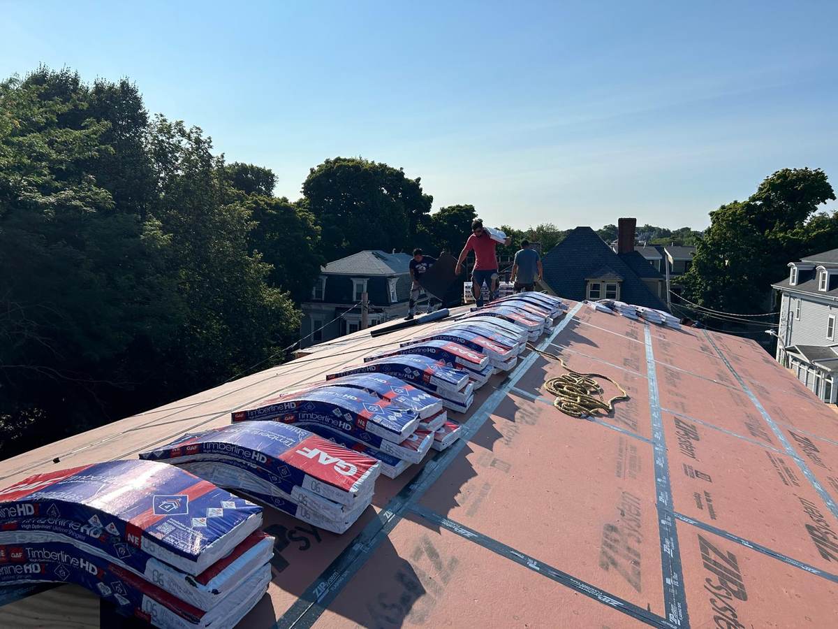 Bundles of asphalt shingles staged on a roof over synthetic underlayment, roofers at the ridge