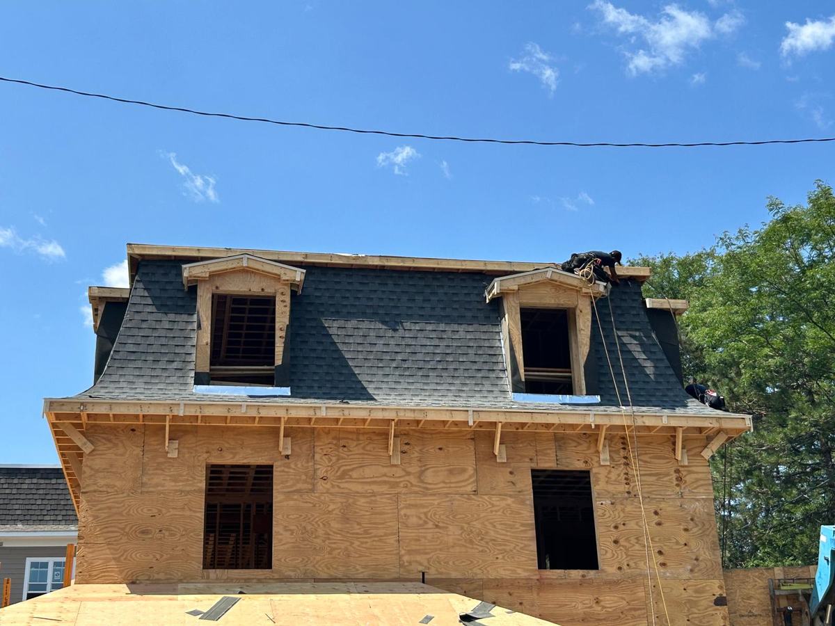 Newly framed house with a gray shingle mansard roof and two dormers, roofers on top