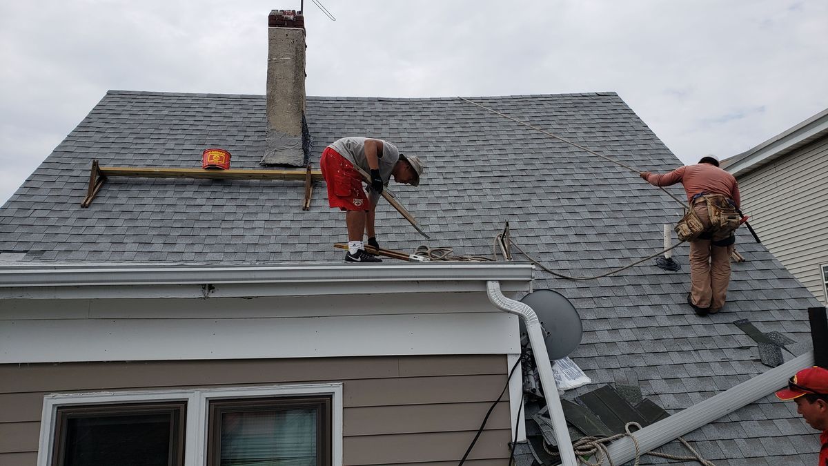 Two roofers working at the eave of a gray shingle roof beside a chimney