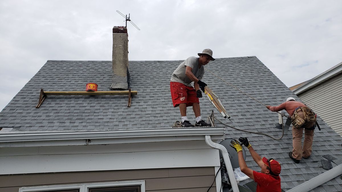 Three roofers installing a gutter at the eave of a gray asphalt shingle roof next to a chimney