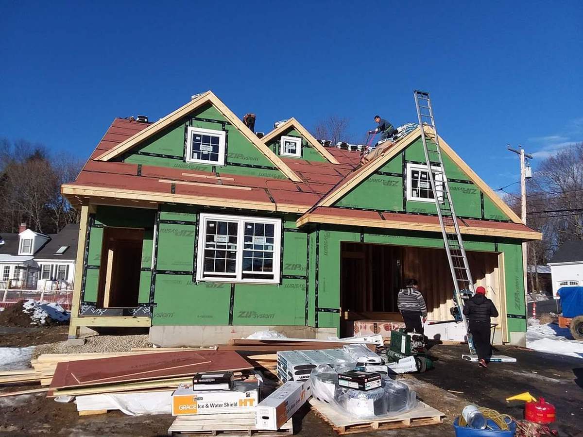 New house with green sheathing and roofers installing shingles under a blue sky